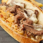 Strip steak sandwich with melted cheese, caramelized onions, and bacon-horseradish spread on a toasted hoagie roll, served with fries in the background.