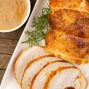 A partially carved boneless, stuffed turkey breast with on a white oblong platter with rosemary garnish, a bowl of apple cider gravy to the left.