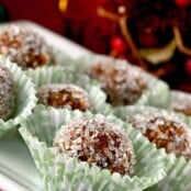Homemade sugar plums rolled in coarse sugar, arranged holiday paper candy cups, Christmas decor in the background.