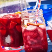 A mason jar filled with summer berry sangria with a red and white striped straw in it, a pitcher with more sangria with fruit in it in the background.
