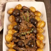 A serving platter of sliced slow-cooker pot roast surrounded by braised mushroom caps and baby potatoes, garnished with a sprig of fresh thyme.