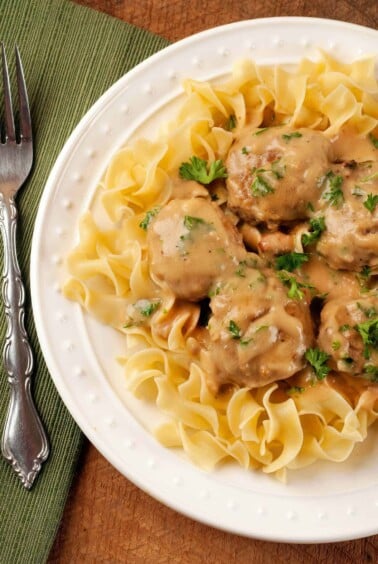 An overhead view of a plate of Swedish meatballs served over egg noodles and garnished with chopped parsley, a napkin and fork to the left.