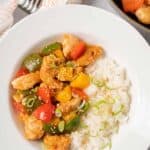 A bowl of sweet and sour chicken with pineapple and red, green, and yellow bell peppers next to a serving of rice and garnished with sesame seeds and scallions.
