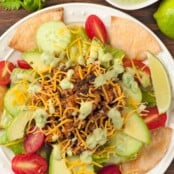 An overhead view of a taco salad on a white plate, surrounded by tortilla chips and topped with shredded cheddar cheese.