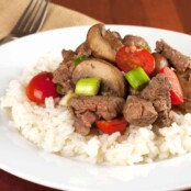 Tenderloin beef tips sautéed with mushrooms, cherry tomatoes, and scallions served over a bed of white rice on a white plate.