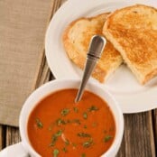A large cup of tomato basil soup garnished with fresh basil next to a plate with a grilled cheese sandwich and a napkin.