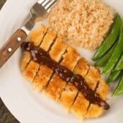 An overhead view a plate with Japanese breaded pork cutlet, sliced and topped with a drizzle of katsu sauce, served with riced cauliflower and sautéed snap peas.