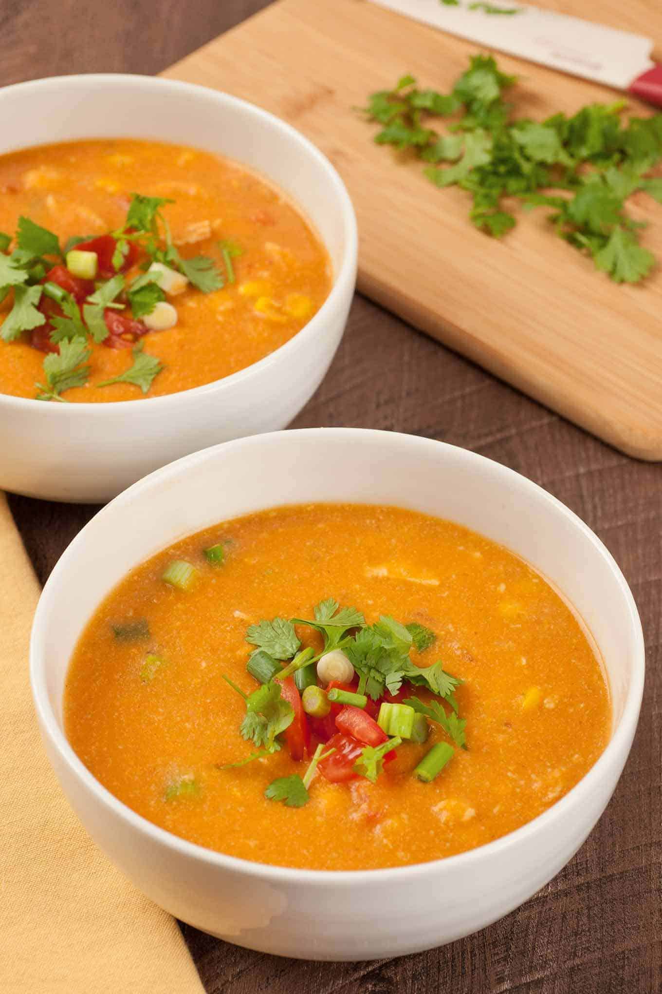 Two bowls of chicken tortilla soup garnished with chopped tomatoes, scallions, and fresh cilantro, with a cutting board topped with cilantro in the background.