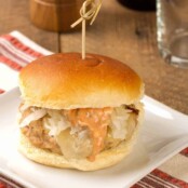 A square white plate atop a striped tea towel with a Reuben-style turkey burger topped with melted Swiss cheese, sauerkraut, and thousand island dressing,