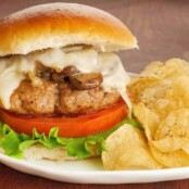 A pan-seared turkey burger topped with sautéed mushrooms and onions and melted muenster cheese, on a bun with lettuce and tomato, and a side of potato chips.