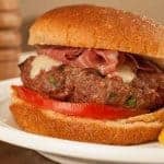 Juicy Tuscan-style burger topped with melted cheese and prosciutto, served on a soft bun with tomato slice and a side of seasoned potato wedges.