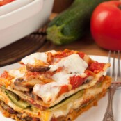 A serving of vegetable lasagna, layered zucchini, eggplant, mushrooms, and cheese, on a plate with a fork, a zucchini, tomato, and casserole of lasagna in the background.