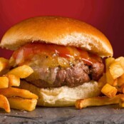 A close view of a grilled burger topped with cheddar cheese, maple ketchup and grilled onions on a slate board, fries to the side.