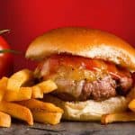 A close view of a grilled burger topped with cheddar cheese, maple ketchup and grilled onions on a slate board, fries to the side.