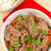 A serving bowl of stir-fried garlic and black pepper beef with strips of red bell pepper and sliced onion, and a garnish of cilantro leaves, a bowl of white rice for serving.