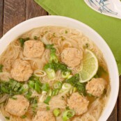 An overhead view of a bowl of soup with thin rice noodles and Vietnamese chicken meatballs, garnished with chopped scallions, cilantro, and lime slices.
