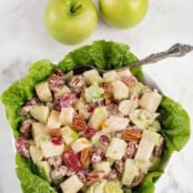 A lettuce lined bowl of Waldorf salad made with crisp apples, fresh orange zest, dried cranberries and toasted pecans, two Granny Smith apples in the background.