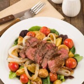 A plate with a salad of grilled sirloin, baby potatoes, cherry tomatoes and mushrooms over mixed greens, napkin with fork and knife to the side.