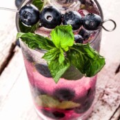 A highball glass filled with a blueberry mojito, muddled berries at the bottom, and a skewer of blueberries and mint sprig as garnish.