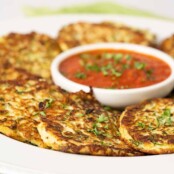 A serving platter of freshly made zucchini fritters garnished with chopped parsley and arranged around a small round bowl of marinara dipping sauce.